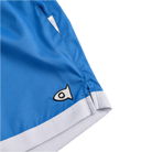 Close-up of blue athletic shorts with a white hem stripe and a small black-and-white fish logo patch, shown on a white background.