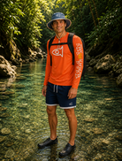 A man standing in a clear, rocky stream surrounded by lush green forest, wearing an orange long-sleeve performance shirt with a white fish logo on the chest and sleeve, navy blue shorts, a blue bucket hat, and a black backpack.