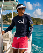 A man wearing a navy long-sleeve performance shirt with a white fish logo, bright pink swim trunks, and a white visor, standing on a boat with blue water and a tropical island in the background.